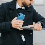 Crop attentive ethnic male office worker in formal apparel with smartphone and laptop checking time on wristwatch in city