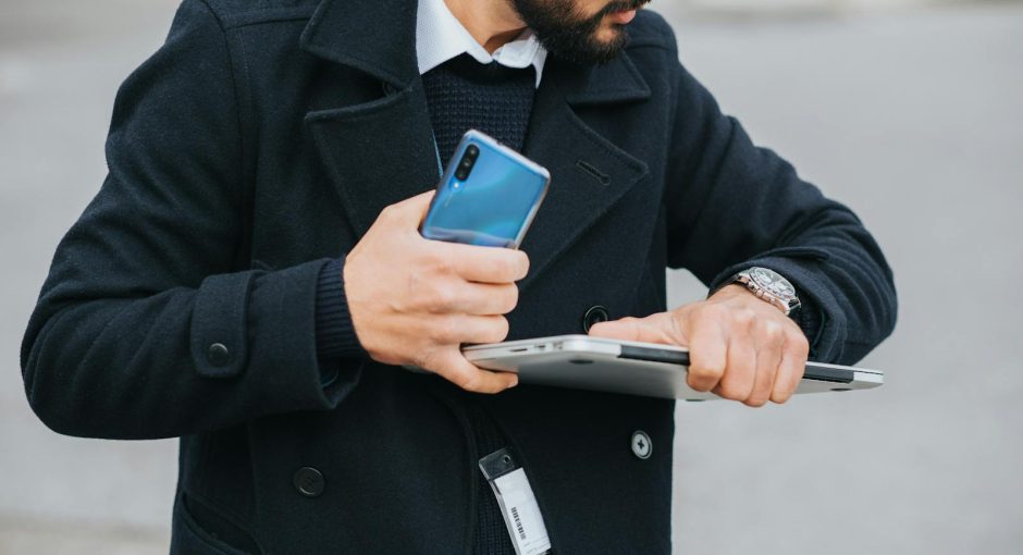 Crop attentive ethnic male office worker in formal apparel with smartphone and laptop checking time on wristwatch in city